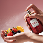 Hand holding a 'Berry Blush' perfume bottle with a spray being applied to a hand holding fruits.