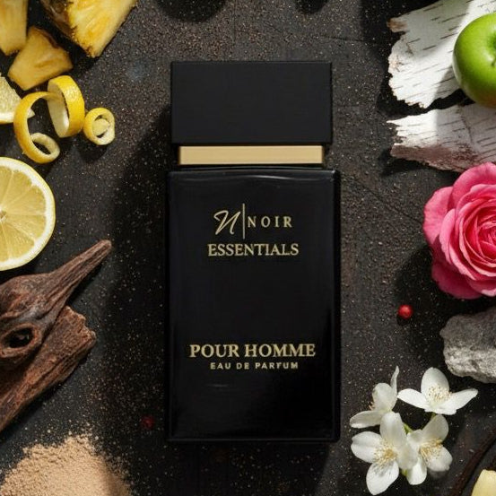 Black perfume bottle labeled 'Noir Essentials Pour Homme' on a dark surface with floral and fruit decorations.
