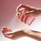 Hand holding a pink perfume bottle spraying onto another hand against a soft pink background